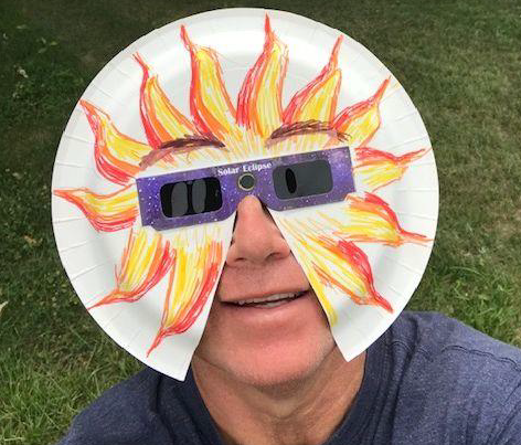 Person wearing decorated paper plate shield with eclipse glasses.