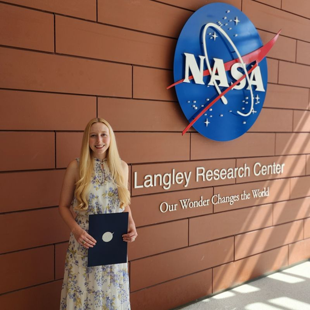 Blond young woman holding an award with NASA logo next to a wall with a large NASA logo above raised lettering that reads Langley Research Center Our Wonder Changes the World